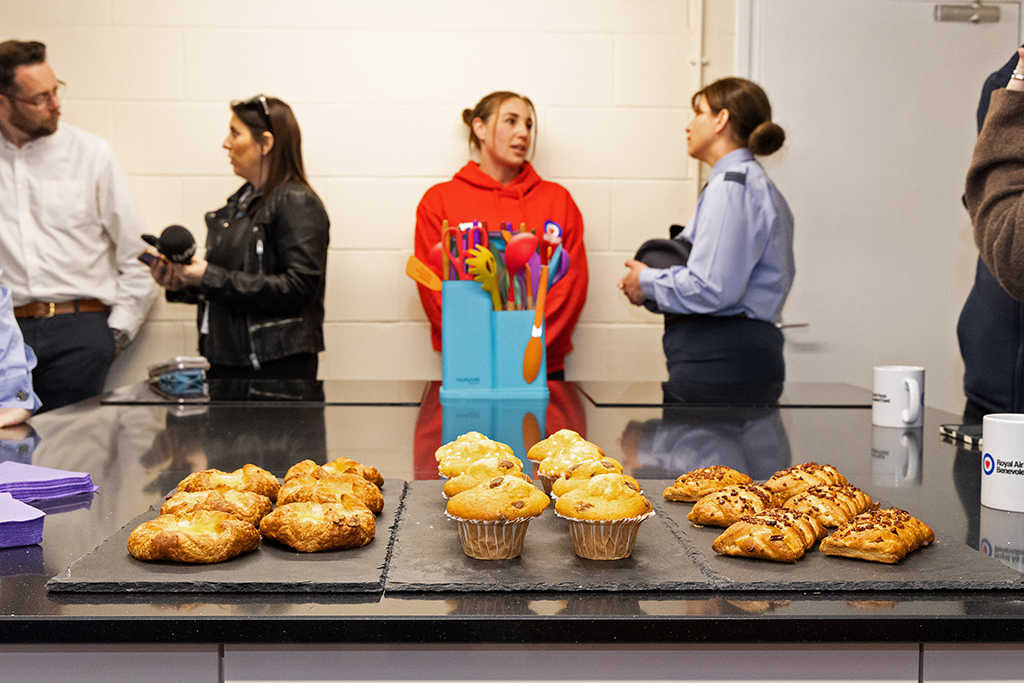 In the Kitchen, cakes, pasties mugs and other fare are displayed in the foreground. Squadron Leader Lucy and invited guests stand in the background.